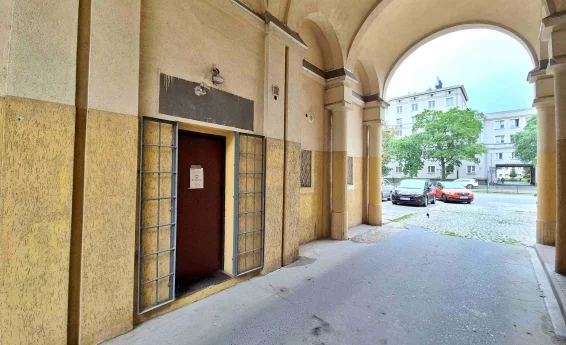 Wejście do lokalu usługowego w arkadach budynku z otwartymi metalowymi kratami oraz widokiem na ulicę i zaparkowane samochody.