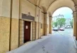 Wejście do lokalu usługowego w arkadach budynku z otwartymi metalowymi kratami oraz widokiem na ulicę i zaparkowane samochody.