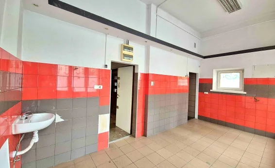 Przestronne pomieszczenie lokalu usługowego z umywalką, czerwono-grafitową okładziną ścienną, drzwiami do kolejnych pomieszczeń oraz niewielkim oknem.